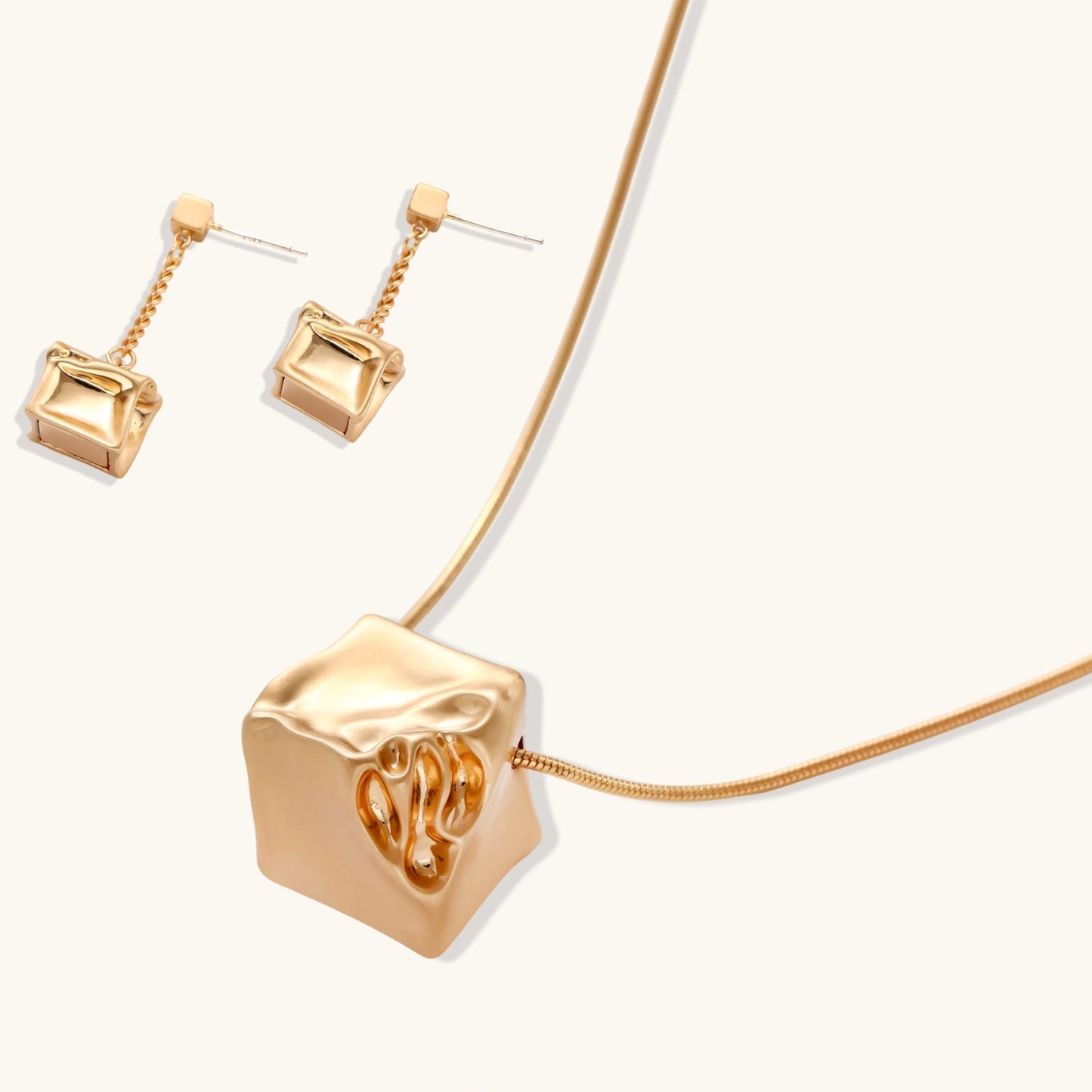 Modern geometric jewelry set featuring crumpled cube pendants in warm gold finish with chain details on beige background.