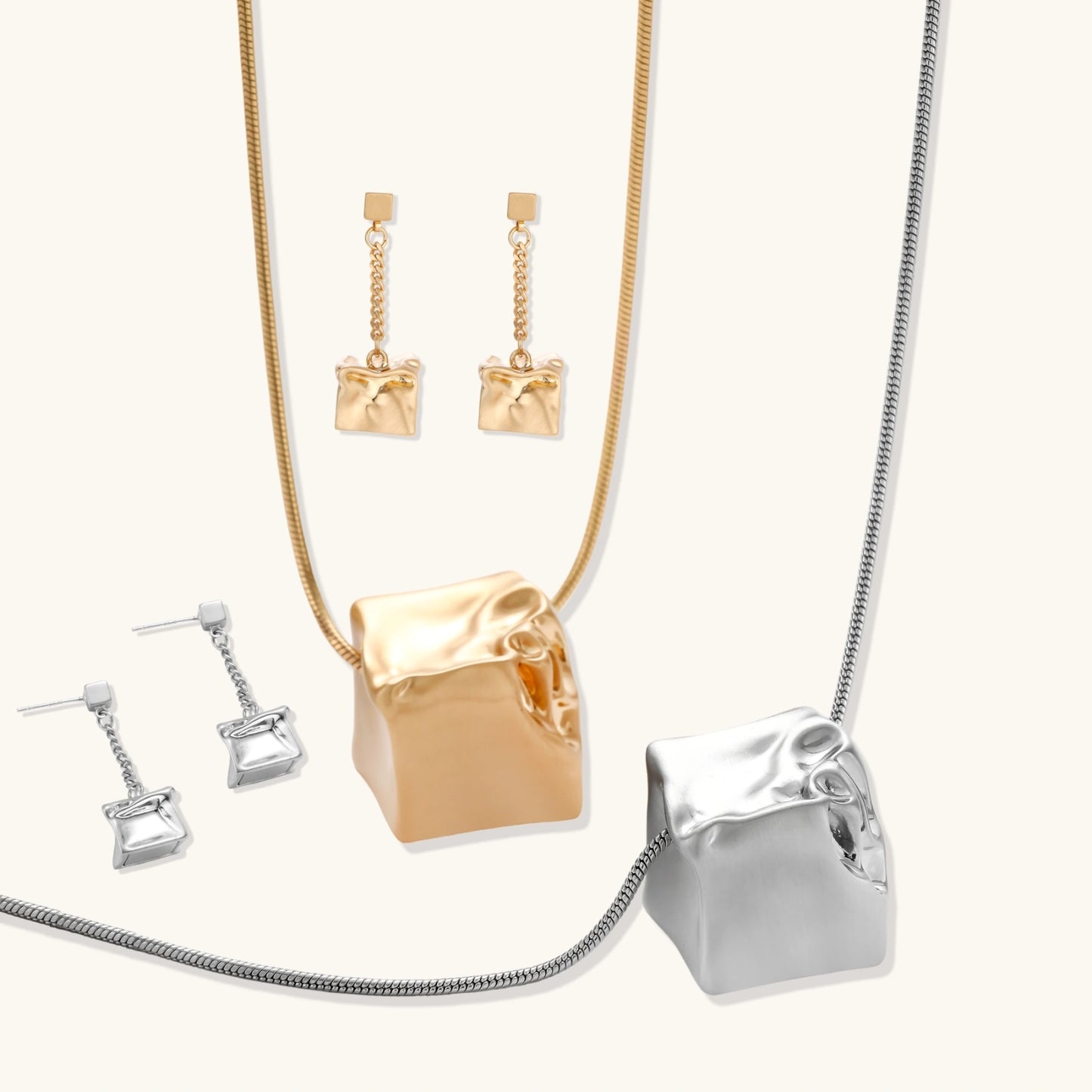 Modern geometric jewelry set featuring crumpled cube pendants in warm gold finish with chain details on beige background.